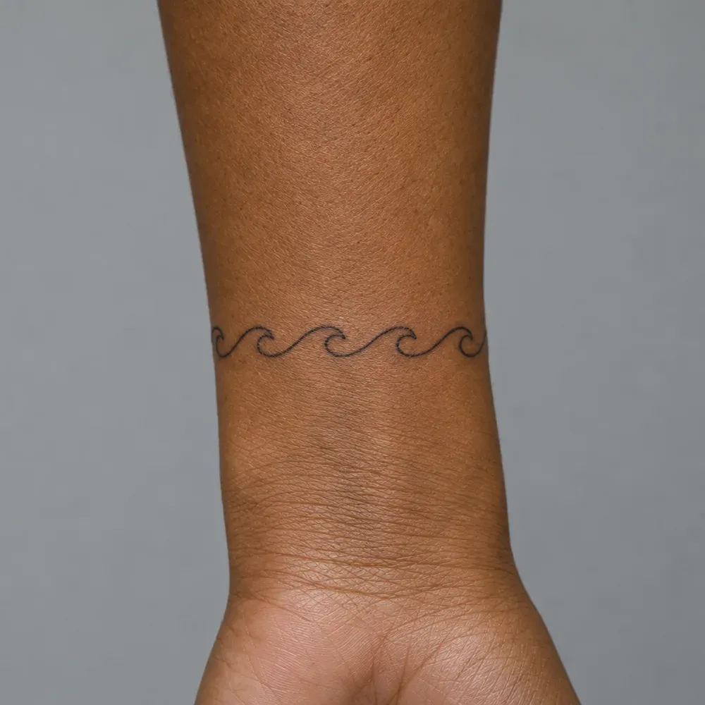 A small fine-line wave band tattooed around the wrist of warm brown skin.