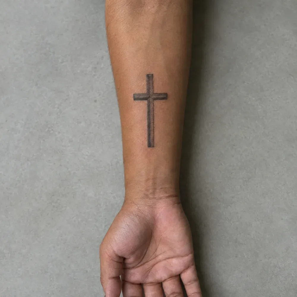 A plain cross tattooed on the inner forearm, shaded down one side of each beam, on warm brown skin with the hand resting palm up.