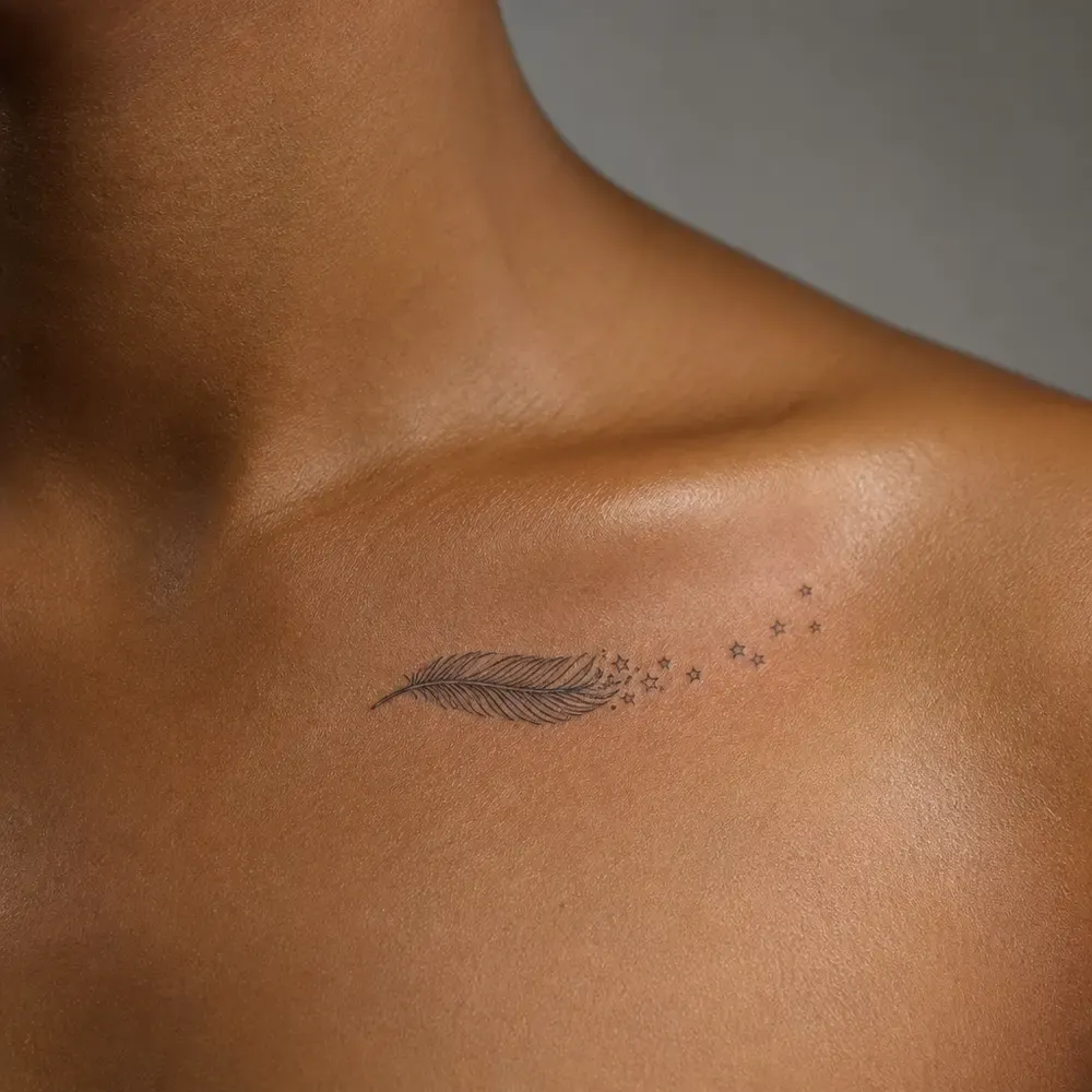 A small fine-line feather trailing tiny stars toward the shoulder tattooed just below the collarbone on warm golden brown skin.