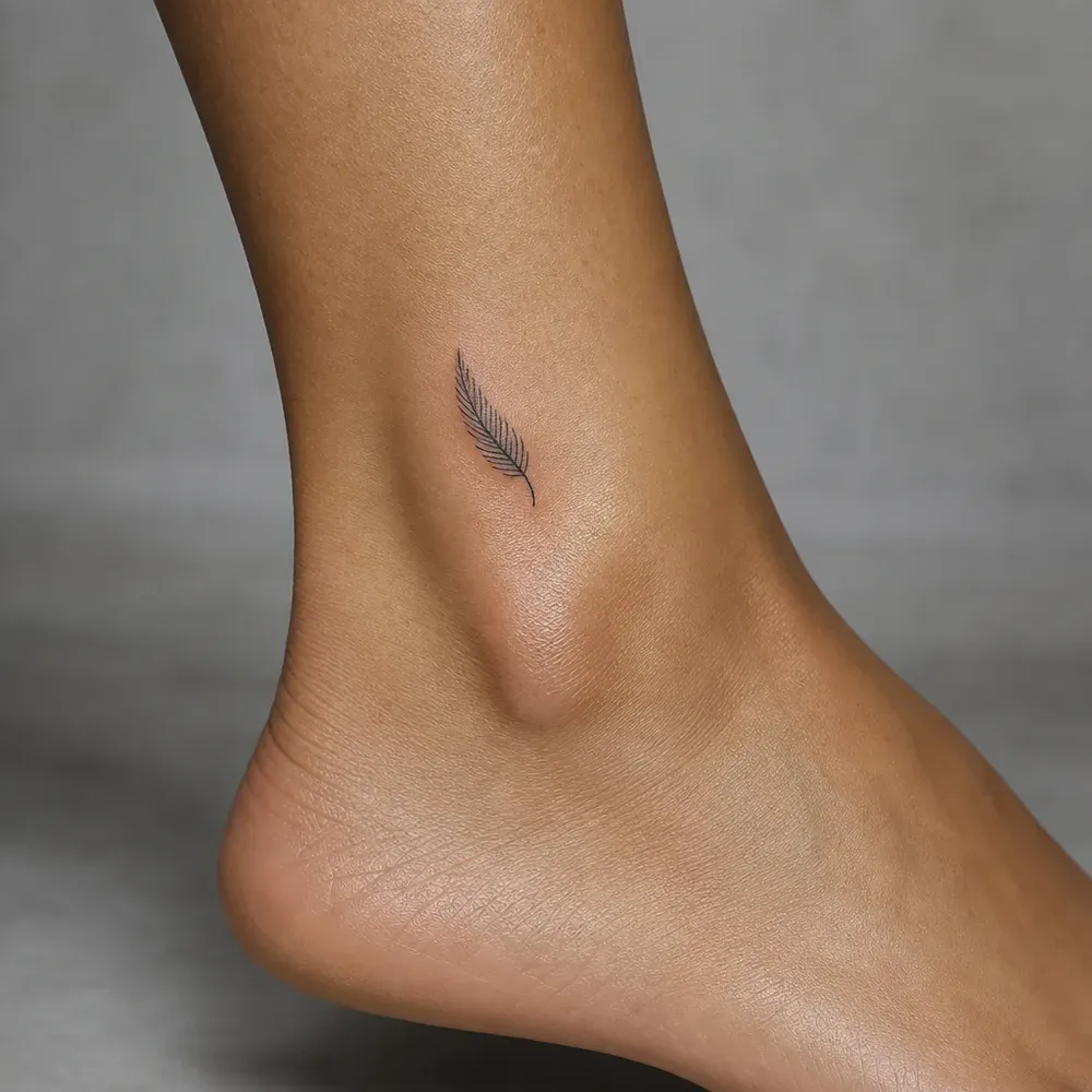 A tiny fine-line feather tattooed above the ankle bone on light tan skin, the bare heel in frame.