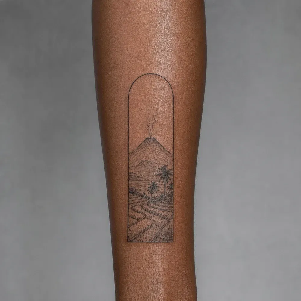 A smoking cone volcano over rice terraces and palms, framed in a tall arch on an inner forearm with deep brown skin.