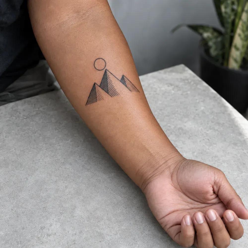 The three hatched peaks and their circle tattooed small on the outer forearm, on warm brown skin, the arm resting on a stone surface.