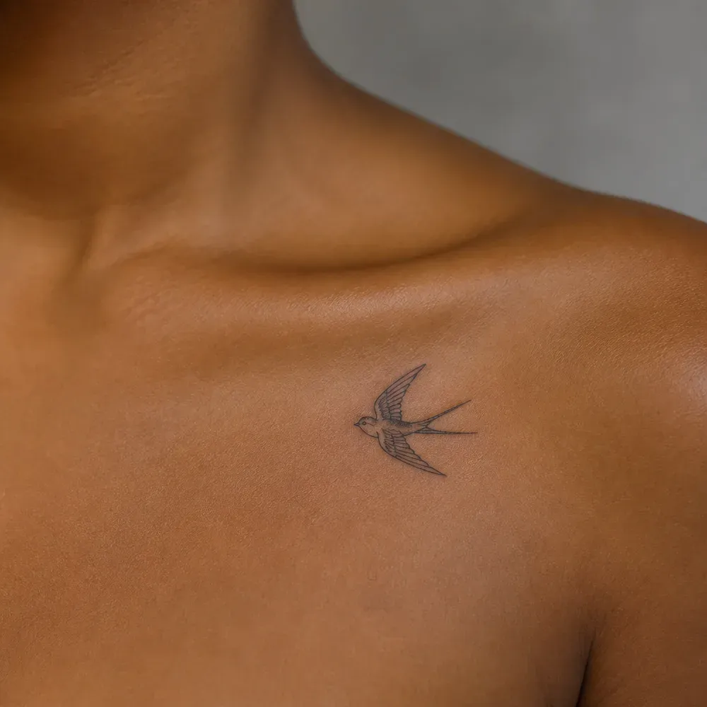 A small fine-line swallow tattooed just below the collarbone on warm brown skin.