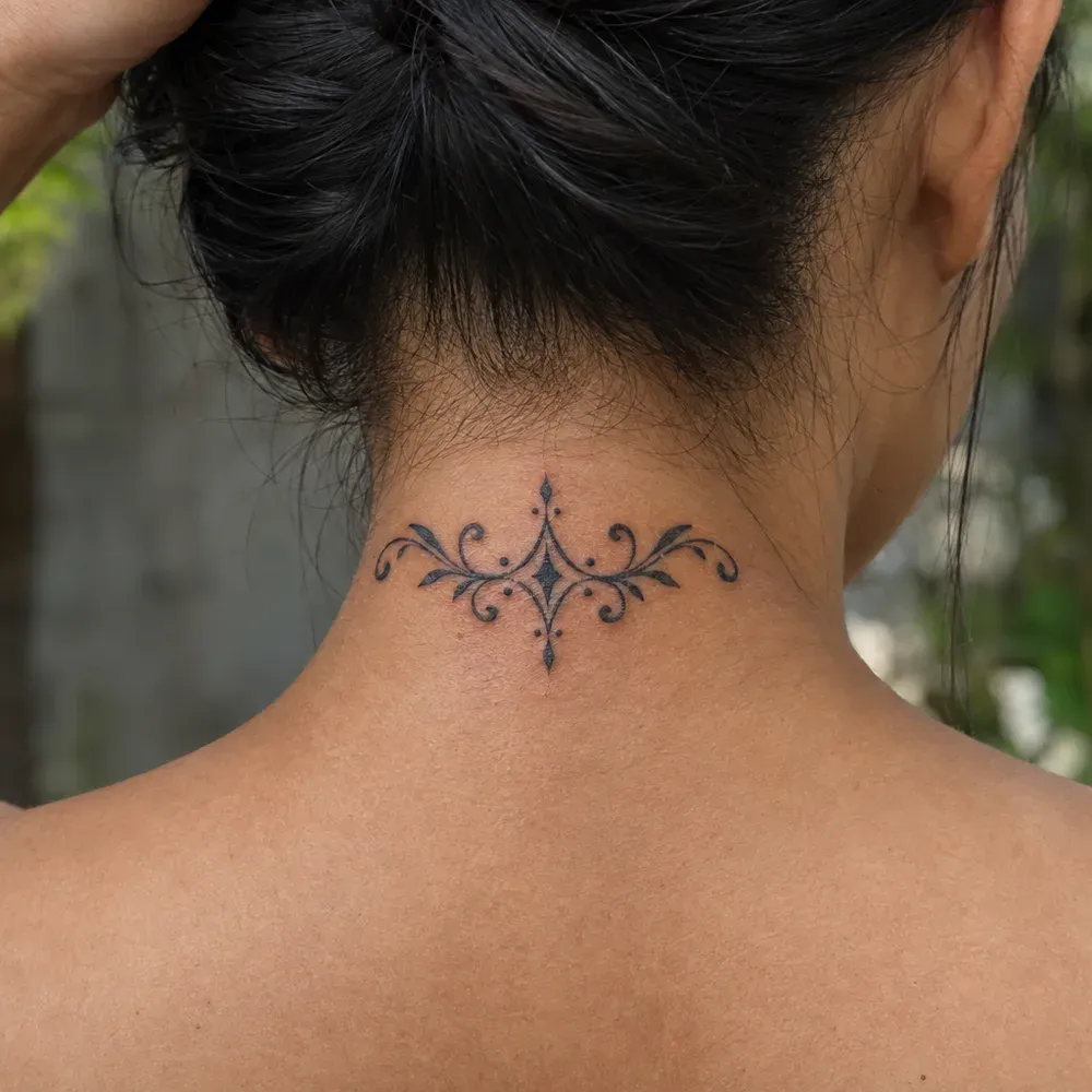 The scrolling nape ornament tattooed across the back of a neck on warm brown skin, the hair lifted and held up out of the way, seen from behind.