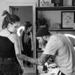 Tattoo artist concentrates on creating a detailed design on a client's arm in a studio setting.